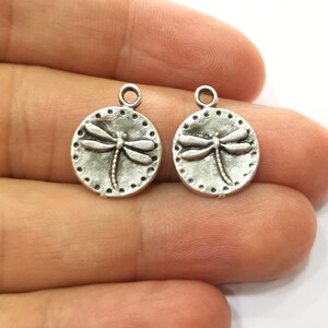 May include: Two silver-toned metal charms in the shape of a circle with a dragonfly design. The dragonfly is raised and has a detailed design.