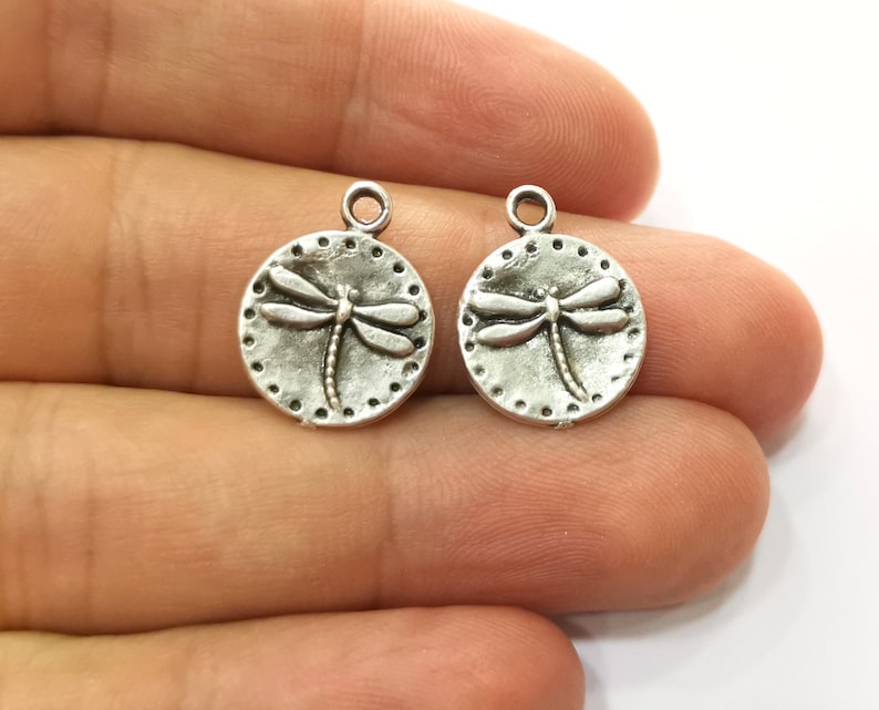 May include: Two silver-toned metal charms in the shape of a circle with a dragonfly design. The dragonfly is raised and has a detailed design.