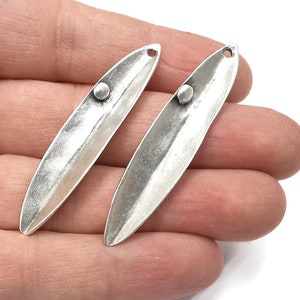 May include: Two silver-toned metal pendant charms shaped like elongated ovals with a rounded edge. Each charm has a small round hole at the top and a small round bead in the center.