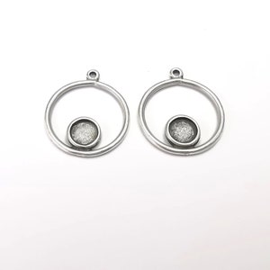 May include: Two silver-coloured metal pendant blanks. Each has a circular frame with a small, round setting for a cabochon or other embellishment. The pendants have a loop at the top for attaching to a chain or other jewellery component.