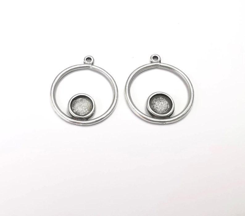 May include: Two silver-coloured metal pendant blanks. Each has a circular frame with a small, round setting for a cabochon or other embellishment. The pendants have a loop at the top for attaching to a chain or other jewellery component.