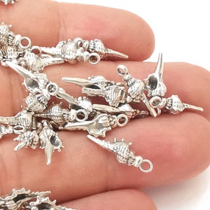 Puede incluir: Una colección de pequeños colgantes de conchas marinas de color plateado. Cada colgante está intrincadamente detallado, pareciendo conchas marinas en miniatura. Tienen un lazo en un extremo para sujetarlos a joyas u otras manualidades. Los colgantes están dispersos sobre un fondo blanco.