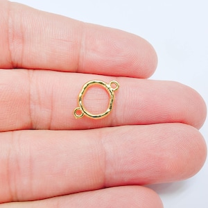 Pode incluir: Um pequeno conector de joalharia em metal dourado com uma forma circular orgânica e irregular e uma superfície texturizada. O componente inclui dois pequenos anéis circulares que se estendem em lados opostos para fixação.