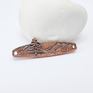 May include: A copper-colored metal pendant with a mountain and tree landscape design. The pendant has a rectangular shape with rounded edges and two small holes. The design includes a detailed depiction of a pine tree, mountains, and a forest.