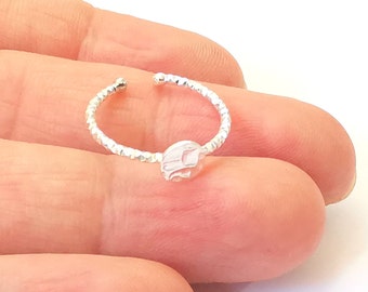 Puede incluir: Un anillo plateado con una banda texturizada y un pequeño diseño estilizado. El anillo es abierto, lo que permite ajustar la talla. El diseño parece ser una pequeña forma abstracta. El anillo se muestra en un dedo.