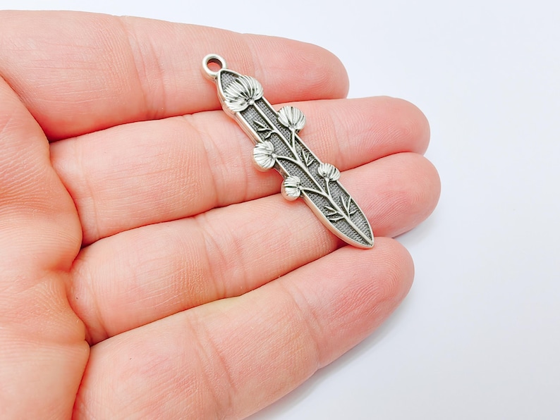 Op de afbeelding: Een zilverkleurige hanger met een bloemmotief. De hanger is langwerpig met een afgeronde bovenkant en heeft een centrale stengel met gedetailleerde bladeren en bloemknoppen. De hanger wordt in een hand gehouden tegen een witte achtergrond.