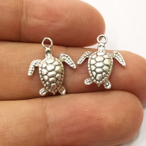 Könnte beinhalten: Zwei silberne Schildkröten-Anhänger mit einem detaillierten Panzerdesign. Die Anhänger haben eine Öse oben zum Anbringen an Schmuck oder andere Bastelarbeiten.