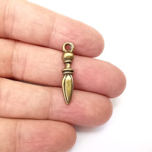 May include: A gold-toned metal charm shaped like a dagger with a decorative top. The charm has a loop at the top for attaching to a necklace or bracelet.