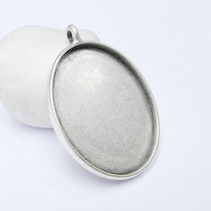 Puede incluir: Base de colgante ovalada plateada con un lazo para una cadena. El colgante tiene una superficie texturizada y un borde elevado para sujetar un inserto. El colgante se muestra sobre un fondo blanco. Mide unos 4 cm de largo.