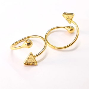 May include: Two gold-colored rings with a unique design. Each ring features a curved band with a spherical accent and a triangular shape at the ends. The rings are displayed on a white surface.