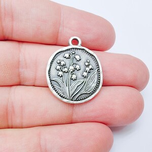May include: A round, silver-colored charm with a detailed embossed design of lily of the valley flowers and leaves. The charm has a small loop at the top for attaching to a necklace or bracelet. The edges are textured.