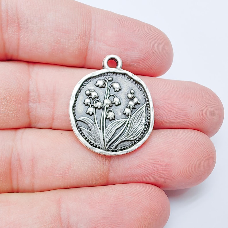 May include: A round, silver-colored charm with a detailed embossed design of lily of the valley flowers and leaves. The charm has a small loop at the top for attaching to a necklace or bracelet. The edges are textured.