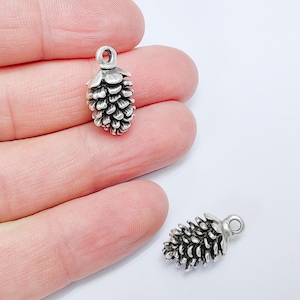 Może przedstawiać: Dwa srebrne charms w kształcie szyszek z pętelką do przymocowania do biżuterii lub rękodzieła.