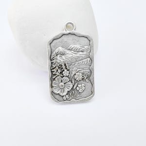 May include: Silver charm featuring an embossed design of a mountain range and wildflowers.