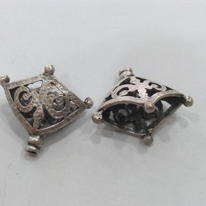 May include: Two silver-toned metal pendant caps with intricate, openwork designs. The caps are shaped like a diamond with rounded corners and have a textured surface.