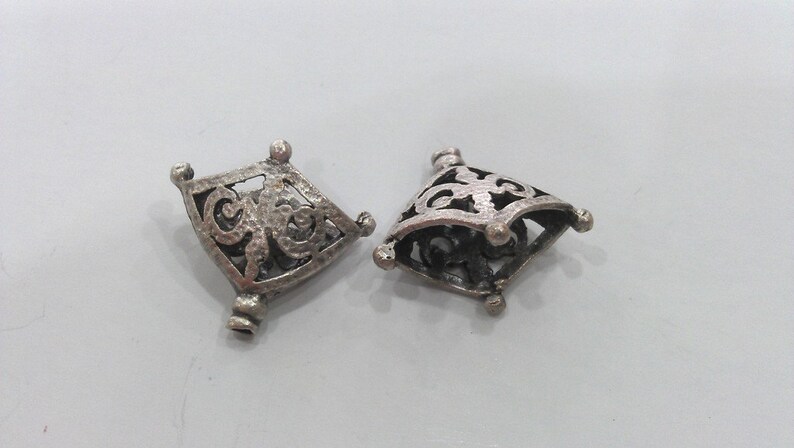 May include: Two silver-toned metal pendant caps with intricate, openwork designs. The caps are shaped like a diamond with rounded corners and have a textured surface.