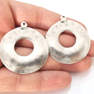 Puede incluir: Dos colgantes de metal de color plateado en forma de círculo con un agujero en el centro. Los colgantes tienen una textura martillada y un borde ligeramente elevado.