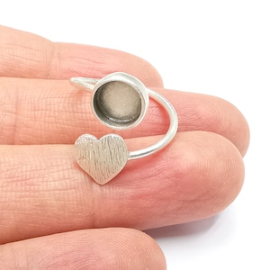 May include: A silver ring with a heart-shaped design and a round bezel setting. The ring is adjustable and can be worn on any finger.