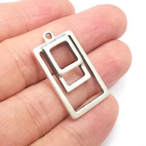 Peut inclure: Pendentif argenté avec un cadre rectangulaire et deux cadres carrés imbriqués. Le pendentif est doté d'une petite boucle en haut pour l'attacher à une chaîne. Le métal présente un aspect légèrement vieilli.
