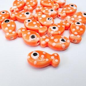 May include: Bright orange fish-shaped beads with white polka dots and black eyes. These ceramic beads have a hole for stringing, ideal for jewellery making or craft projects. The beads are a vibrant orange colour.