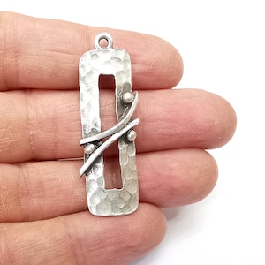 May include: Silver-toned pendant with a rectangular frame and a hammered texture. The frame has a cutout in the center and is decorated with a silver-toned wire design with small beads.