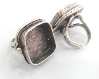 May include: Silver ring with a hinged square compartment. The ring is open and the compartment is empty.