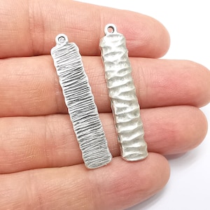 May include: Two silver metal rectangular charms with textured surfaces. The charms have a loop at the top for attaching to jewelry or other crafts.