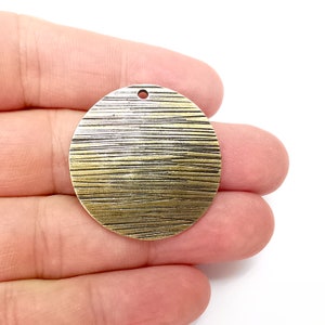 以下が含まれることがあります： ストライプのテクスチャ加工が施された丸い真鍮色の金属製のペンダント。ペンダントの上部に吊り下げ用の穴があります。