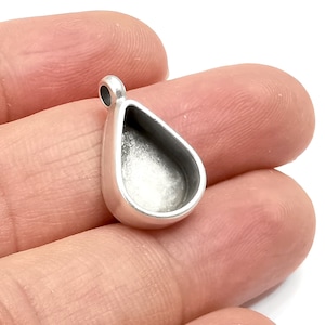 May include: Silver teardrop pendant with a round opening for adding a resin or glass cabochon. The pendant is shown on a finger.