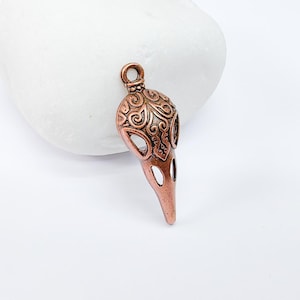 Puede incluir: Un colgante de color cobre con forma de cabeza de pájaro con intrincados diseños en espiral. El colgante tiene un agujero en la parte superior para sujetar una cadena o cordón.