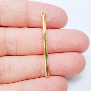 May include: A gold-coloured, slender bar pendant with a small loop at the top for attaching to a chain or other jewellery component. The pendant is held in a person's hand against a white background.