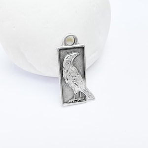 Op de afbeelding: Zilveren bedel met een gedetailleerd ravenontwerp, de vogel is in profiel afgebeeld met zijn vleugels gevouwen, de bedel is rechthoekig van vorm met een glad oppervlak.