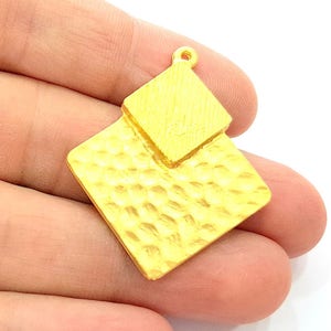 以下が含まれることがあります： 金色のペンダント。テクスチャ加工とハンマー加工が施されています。ペンダントは、対角線のストライプ模様と、くぼみのあるテクスチャの2つの重なり合うダイヤモンド型を特徴としています。上部に吊り下げ用の小さなループが付いています。