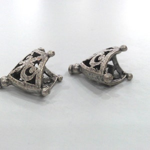 May include: Two silver metal decorative end caps with an intricate design. The end caps are triangular in shape and have a hollow centre.