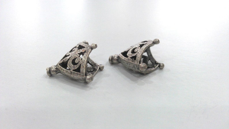 May include: Two silver metal decorative end caps with an intricate design. The end caps are triangular in shape and have a hollow centre.