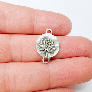 May include: A small, silver-coloured charm featuring a detailed lotus flower design. The charm is circular with a loop at the top and bottom for attaching to jewellery. The lotus flower is in the centre of the charm.