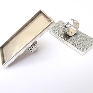 May include: Two silver ring blanks with a rectangular tray for resin or other embellishments. The tray is smooth and the ring blank has a textured design.
