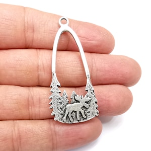 Könnte beinhalten: Silberfarbener Metallanhänger mit einer Elch-Silhouette, die in einer Waldszene steht. Der Anhänger ist oval geformt mit einem Ausschnitt in der Mitte.