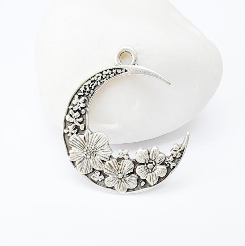May include: Silver-toned crescent moon pendant with floral detailing. The pendant features three large flowers and smaller floral accents along the moon's curve. It has a loop at the top for easy attachment to a chain or cord.