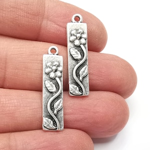 May include: Two silver-toned metal charms with a floral design. Each charm features a stylized flower with leaves and a stem.