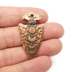 May include: A copper-toned arrowhead pendant with a hammered texture and a design of crescent moons and a circle.