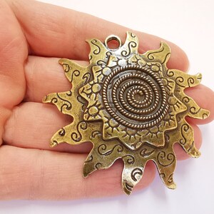 K&ouml;nnte beinhalten: Ein goldfarbener Metall-Sonnenanh&auml;nger mit einem Spiraldesign in der Mitte und filigranen Details an den R&auml;ndern. Der Anh&auml;nger hat eine &Ouml;se oben zum Anbringen an einer Kette oder Schnur.