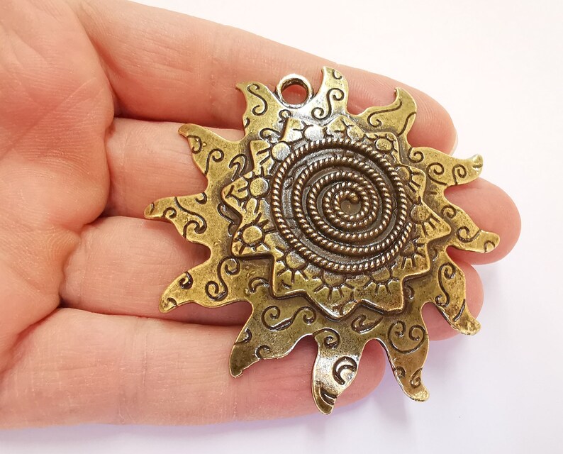 K&ouml;nnte beinhalten: Ein goldfarbener Metall-Sonnenanh&auml;nger mit einem Spiraldesign in der Mitte und filigranen Details an den R&auml;ndern. Der Anh&auml;nger hat eine &Ouml;se oben zum Anbringen an einer Kette oder Schnur.