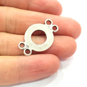 Puede incluir: Conector de metal plateado con dos bucles a cada lado. El conector tiene forma de círculo con un círculo más pequeño cortado en el centro.