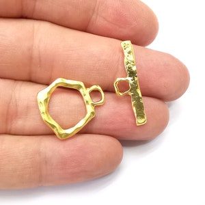 Könnte beinhalten: Goldfarbener Metall-Schiebeverschluss mit strukturierter Oberfläche. Der Verschluss besteht aus einer rechteckigen Stange mit einer Schlaufe an jedem Ende und einer separaten, unregelmäßig achteckigen Schlaufe.