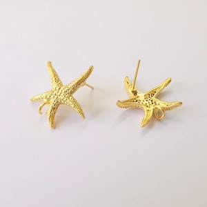 Puede incluir: Un par de pendientes de botón dorados con forma de estrella de mar. Los pendientes tienen superficies texturizadas y cierres de poste. Se muestran sobre un fondo blanco.