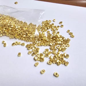 May include: A collection of small, gold-colored metal beads, scattered on a white surface. The beads are oval-shaped with a hole in the center, and some are still in a clear plastic bag. The beads are likely for jewelry making or crafts.