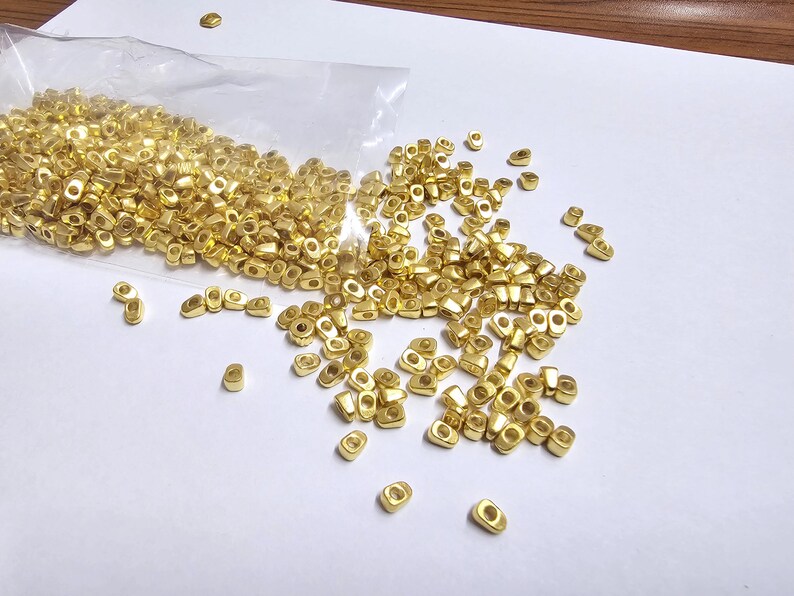 May include: A collection of small, gold-colored metal beads, scattered on a white surface. The beads are oval-shaped with a hole in the center, and some are still in a clear plastic bag. The beads are likely for jewelry making or crafts.