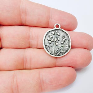 May include: A silver-colored round pendant with a detailed relief of lily of the valley flowers and leaves. The pendant has a small loop at the top for attaching to a chain or cord. The design is set within a textured border.