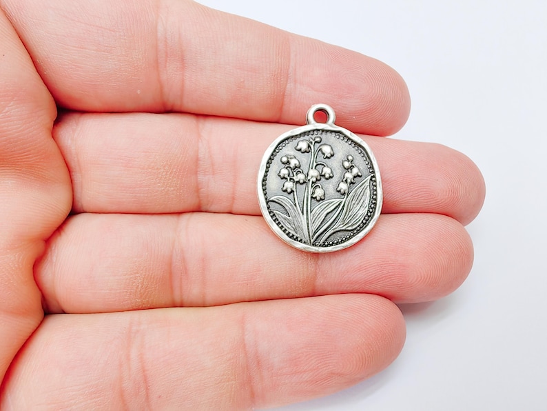 May include: A silver-colored round pendant with a detailed relief of lily of the valley flowers and leaves. The pendant has a small loop at the top for attaching to a chain or cord. The design is set within a textured border.